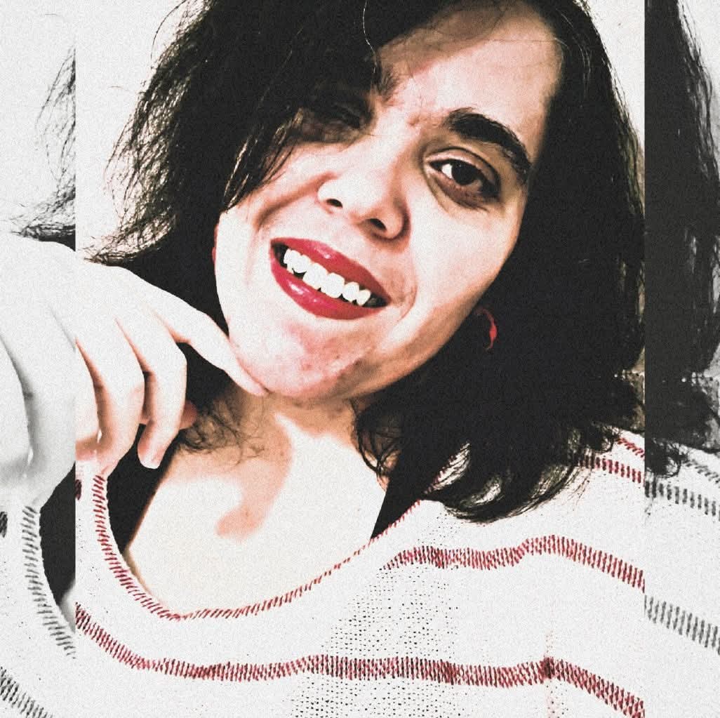 Mulher de pele clara sorrindo em um close, com batom vermelho vivo e cabelo escuro e ondulado caindo sobre parte do rosto. Os dentes estão bem visíveis no sorriso e os olhos castanhos aparecem voltados para a câmera. A iluminação é forte e deixa a pele be